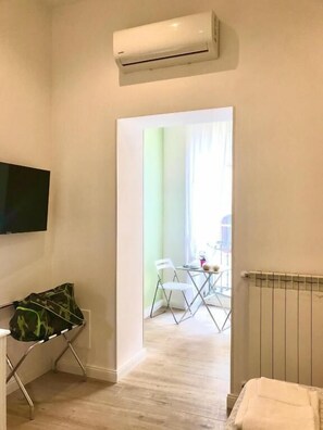 A room with an air conditioner, a TV mounted on the wall, a folding chair, a radiator, and a view through an open doorway to a dining area with a table and chairs. - Agorà (Naples)