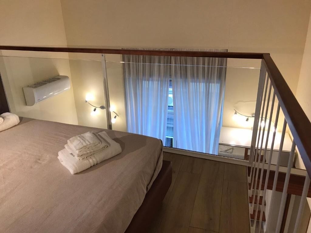 A hotel room with a bed, a glass partition, a view of a building, and a mounted air conditioner.