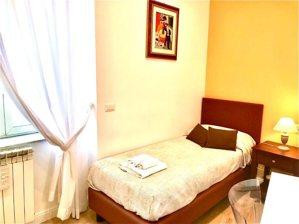 A single bed with a headboard, a bedside table with a lamp, a framed picture on the wall, and a window with curtains.