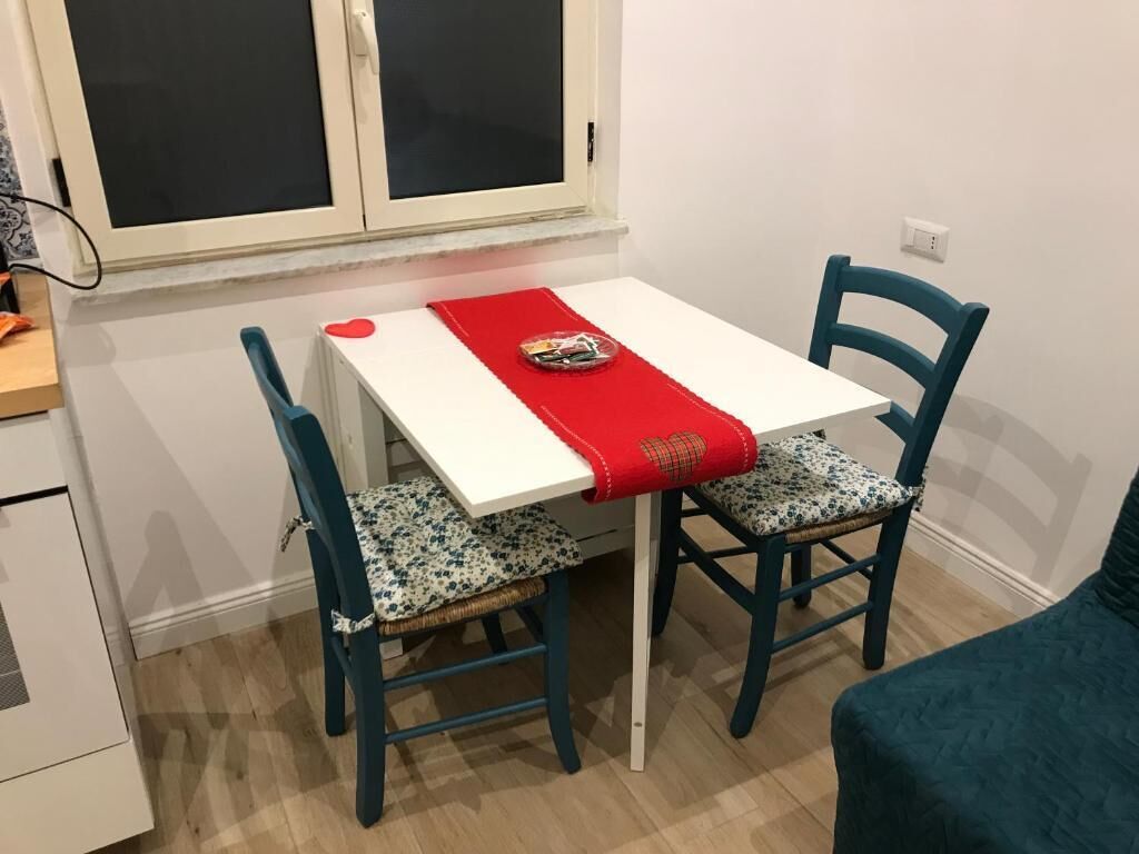 A small dining area with a white table, two blue chairs, a red placemat, and a window above.