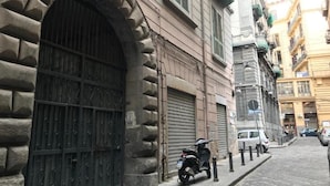 A narrow street with a stone building featuring a large arched entrance and a closed shutter. A scooter is parked on the sidewalk, and a car is visible in the background. - Agorà Rooms (Naples)