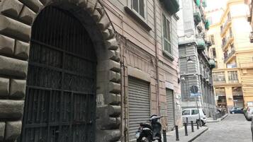 Eine enge Straße mit einem Steinhaus, das einen großen Bogenbogen und einen geschlossenen Rollladen aufweist. Ein Roller steht am Gehweg geparkt, und im Hintergrund ist ein Auto sichtbar.