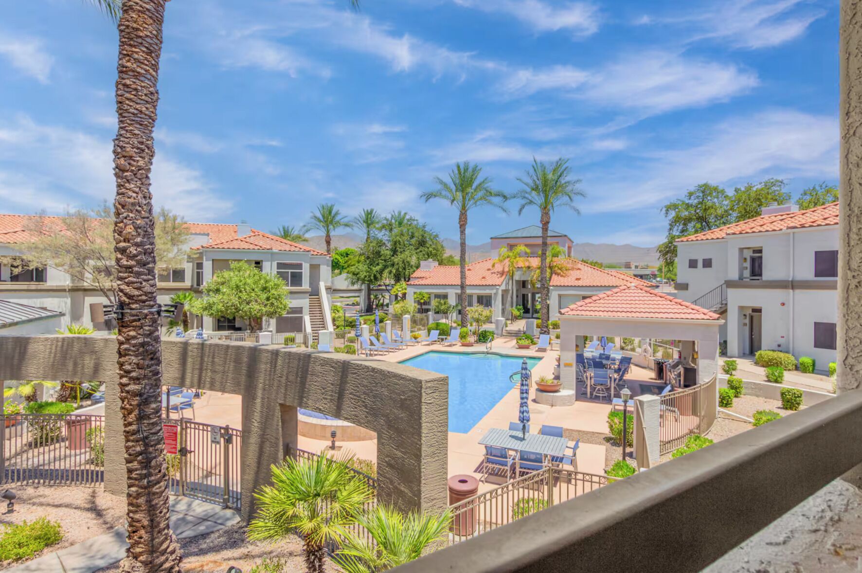 A pool area with lounge chairs, palm trees, and a building with a red-tiled roof.