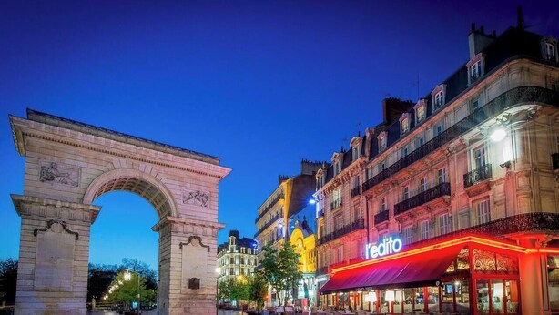 A historic archway with classical architecture, adjacent to a brightly lit building with a red awning and a sign that reads 'Editio.' - 1,001 Wonders (Fénay)