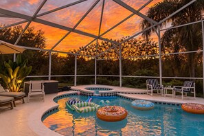Piscina al aire libre con sillas de descanso y flotadores, rodeada por un espacio cubierto con vista a árboles y un atardecer.