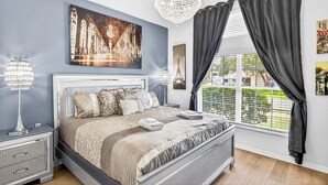 Une chambre à coucher avec un lit, un lustre, une table de chevet, une fenêtre avec des rideaux et un tableau au mur.