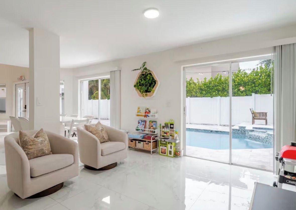 Ein geräumiges Wohnzimmer mit zwei Sofas, einem Couchtisch und einem Bücherregal. Ein großes Fenster bietet Blick auf einen Swimmingpool und Grünflächen.