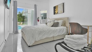 Un dormitorio moderno con una cama grande, un televisor de pantalla plana, un cuadro y vistas a una piscina y edificios.