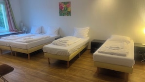A hotel room with three single beds, a wooden floor, a window with curtains, and a painting on the wall.