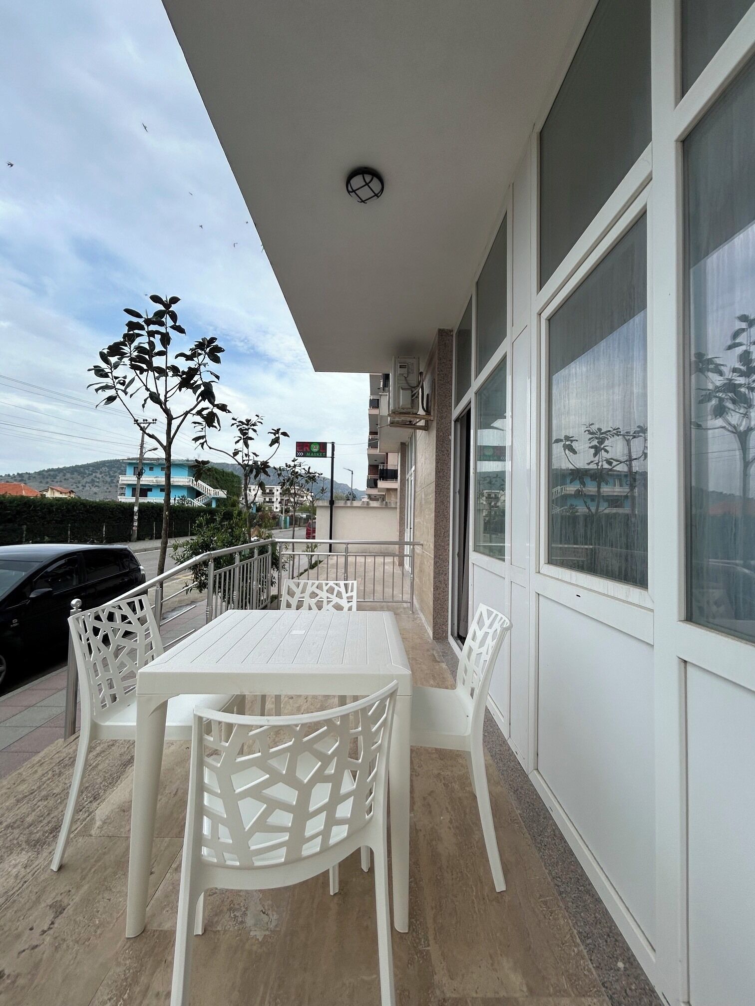 Un balcón con una mesa y sillas blancas, vista a una calle y un edificio con grandes ventanales.