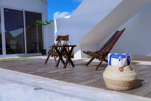 Un espacio de asiento al aire libre con una mesa y sillas, una cesta tejida y un edificio de fachada blanca con grandes ventanales.