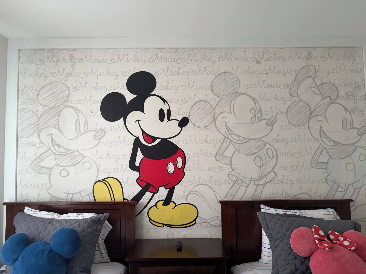 Una habitación con un mural grande de Mickey Mouse en la pared, dos camas con almohadas y una mesita de noche.