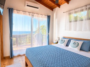 A bedroom with a bed, curtains, a nightstand, and a view of the outdoors. - Terra Chula 1BR Bungalow with Ocean & Nature Views (Roatán)