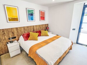 A modern bedroom with a wooden headboard, a white bed with red and yellow pillows, a white nightstand with a remote control, and artwork on the wall. - Sand, Sea and Love by Interhome (Puerto Rico)