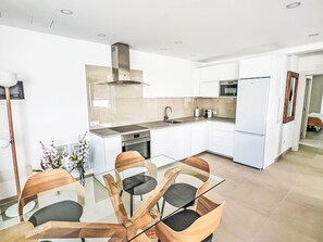 A modern kitchen with white cabinets, a stainless steel range hood, a built-in oven, and a microwave. There is a glass dining table with four chairs. - Sand, Sea and Love by Interhome (Puerto Rico)