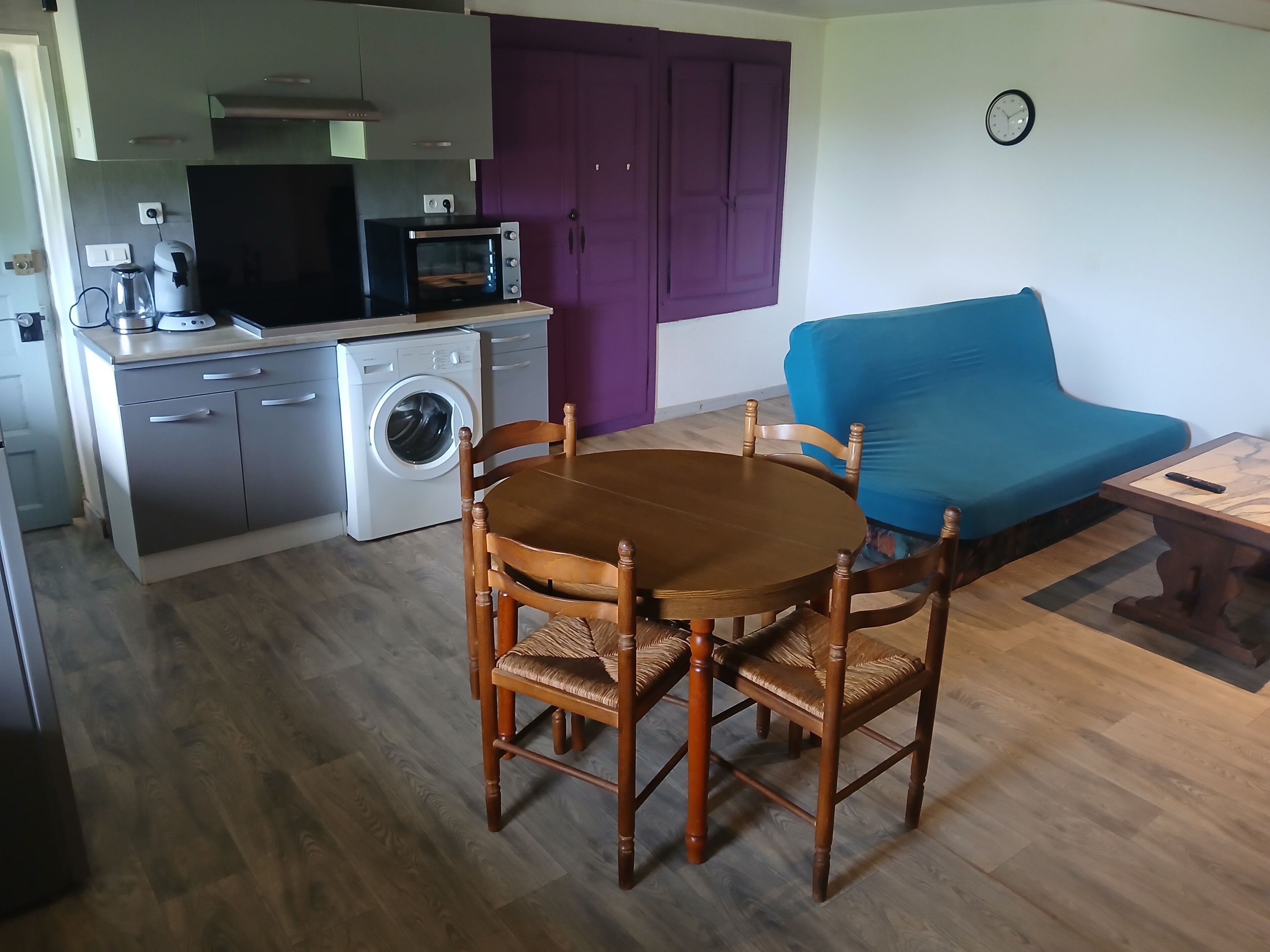 A compact kitchen with a microwave, washing machine, and a purple door. A wooden dining table with chairs is in the foreground.