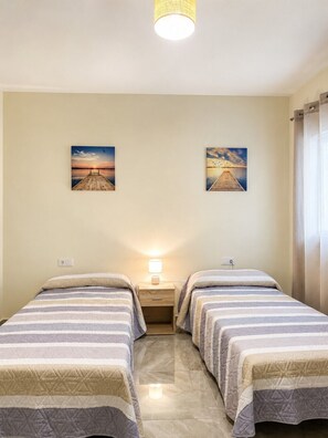 Une chambre d’hôtel avec deux lits, une table de chevet avec une lampe et deux tableaux accrochés au mur représentant un coucher de soleil sur une jetée.