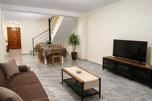Un salon comprenant un canapé, une table basse, un téléviseur posé sur un meuble, une table à manger avec des chaises et un escalier.