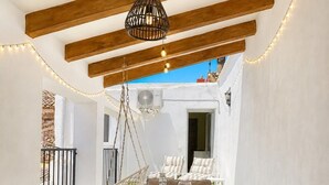 An outdoor dining area with a table set for two, a hanging light fixture, and a view of a garden and building in the background.
