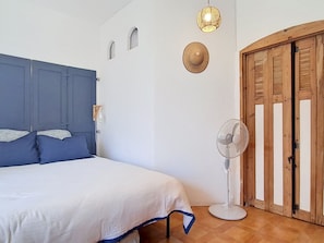 A bedroom with a bed, a standing fan, a wooden door, and a wall-mounted light fixture. - Le Weekend by Interhome (Saint Cyr sur mer Les Lecques)