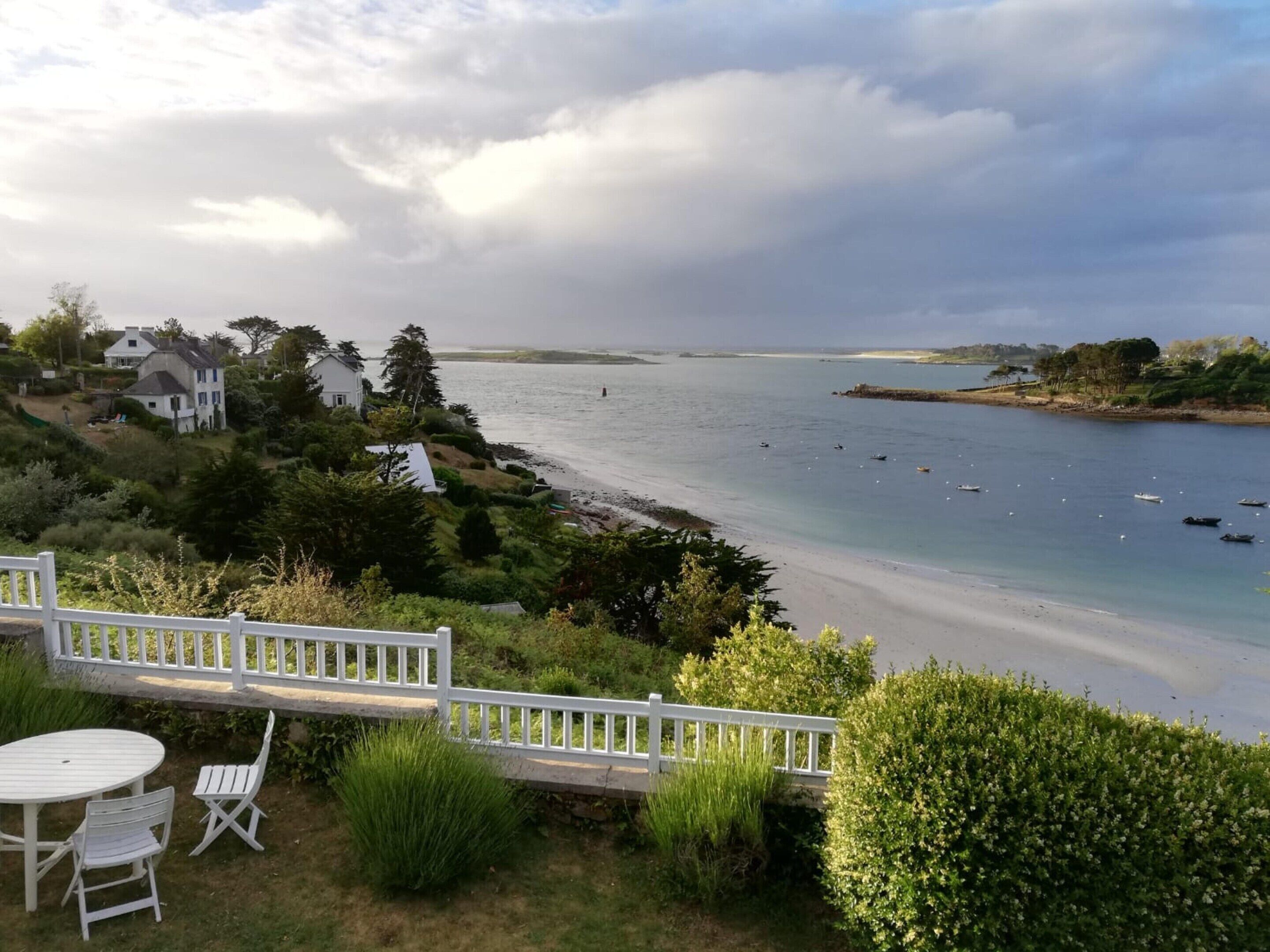 A coastal view with a white fence, a table with chairs, and houses along the shoreline.