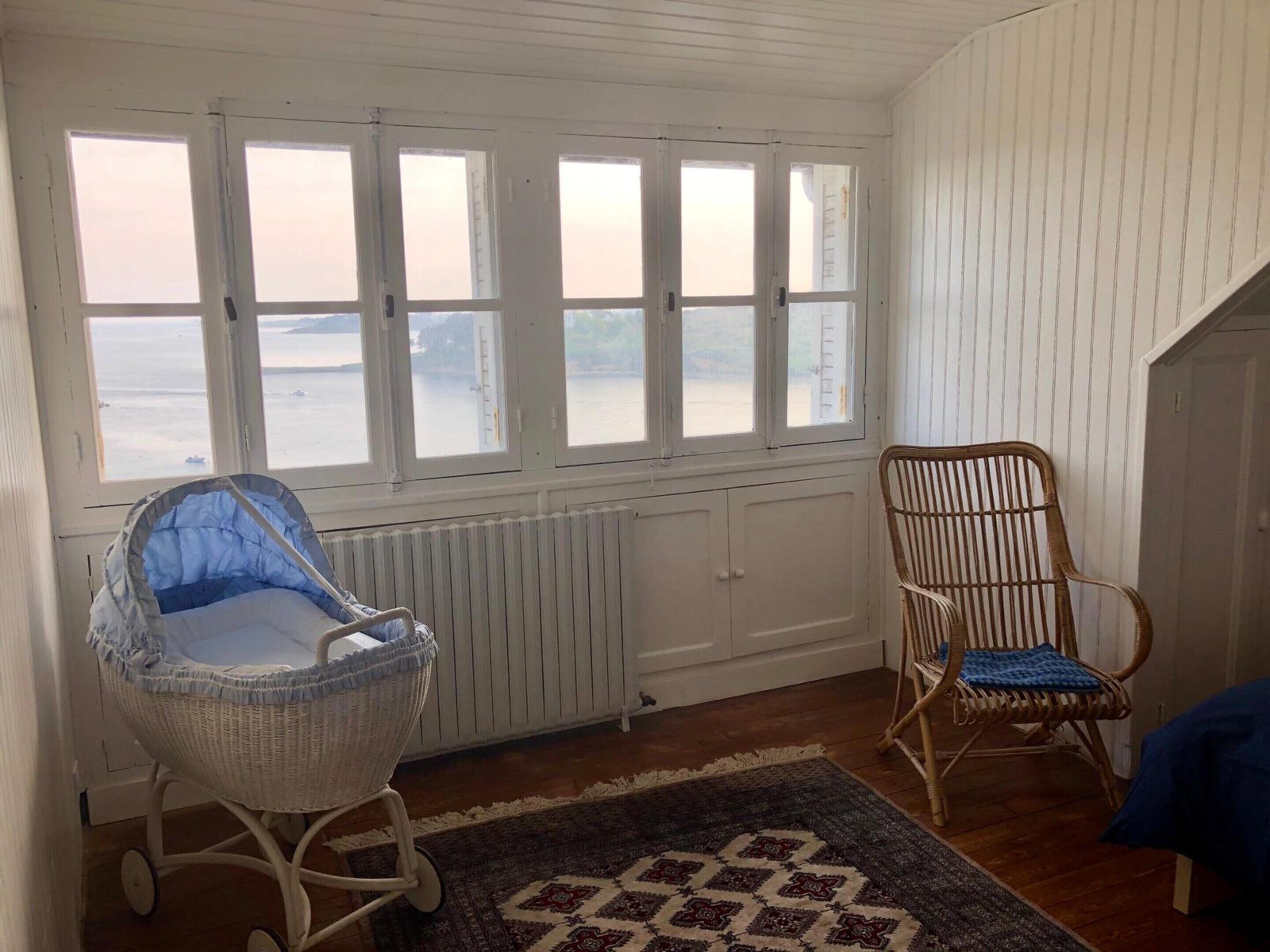 A room with a view of the sea, a wicker baby rocker, a wooden chair, and a patterned rug.