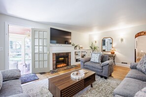 A living room with a fireplace, grey sofas, a coffee table, and a TV mounted above the fireplace. - Brighton Stay Near URMC | King Bed, Home Cinema & Backyard (Rochester)