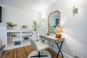 Una habitación con pared blanca, un escritorio de madera con lámpara, una silla, un espejo, una estantería y una planta.