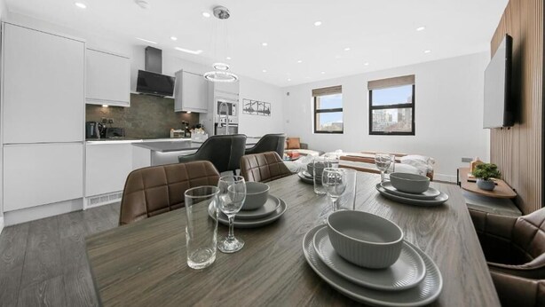 Una cocina moderna con armarios blancos, un comedor con mesa de madera dispuesta para cuatro personas y un conjunto de sillones de cuero marrón.