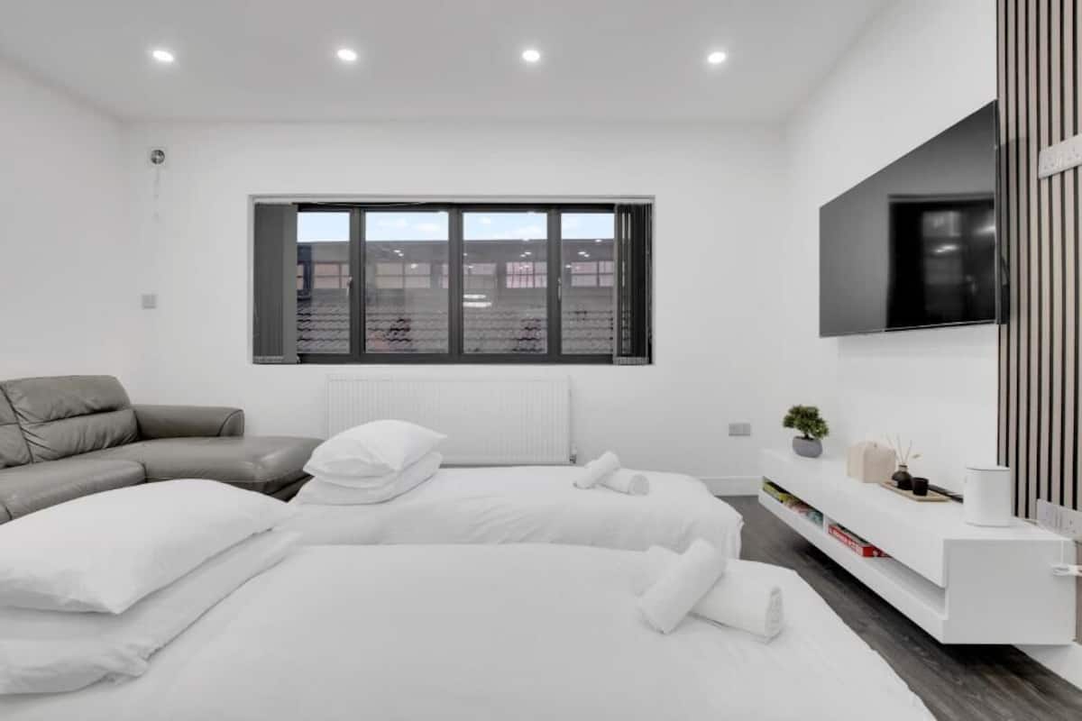 Quarto de hotel moderno com uma janela grande, um sofá, duas camas, uma TV e um console branco.