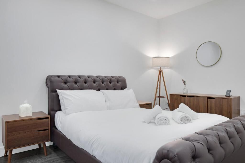 A neatly made bed with white linens, a bedside lamp, a round mirror, and a wooden nightstand with a tissue box.