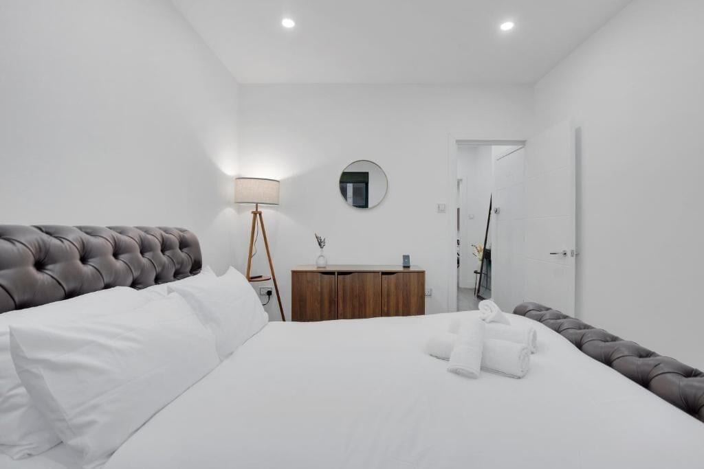 A modern bedroom with a tufted headboard, a wooden dresser, a round mirror, and a bedside lamp.
