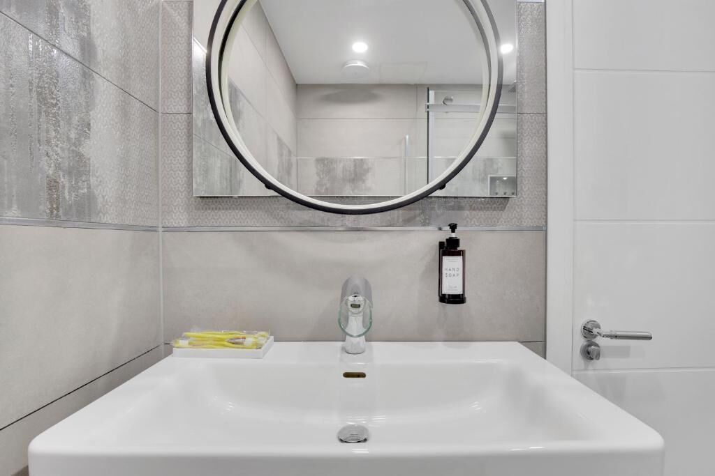 A modern bathroom with a rectangular sink, a round mirror, and a handheld soap dispenser.