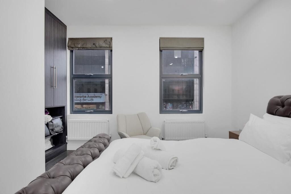 A modern bedroom with a large bed, a seating area, and two windows with blinds.