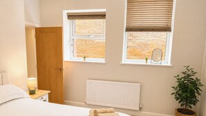 Een slaapkamer met een bed, twee ramen met zonwering, een radiator, een deur, een nachtkastje met een lamp en een potplant.