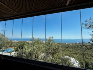 Vista a través de una ventana: cielo despejado, masa de agua distante y paisaje con árboles y un edificio.