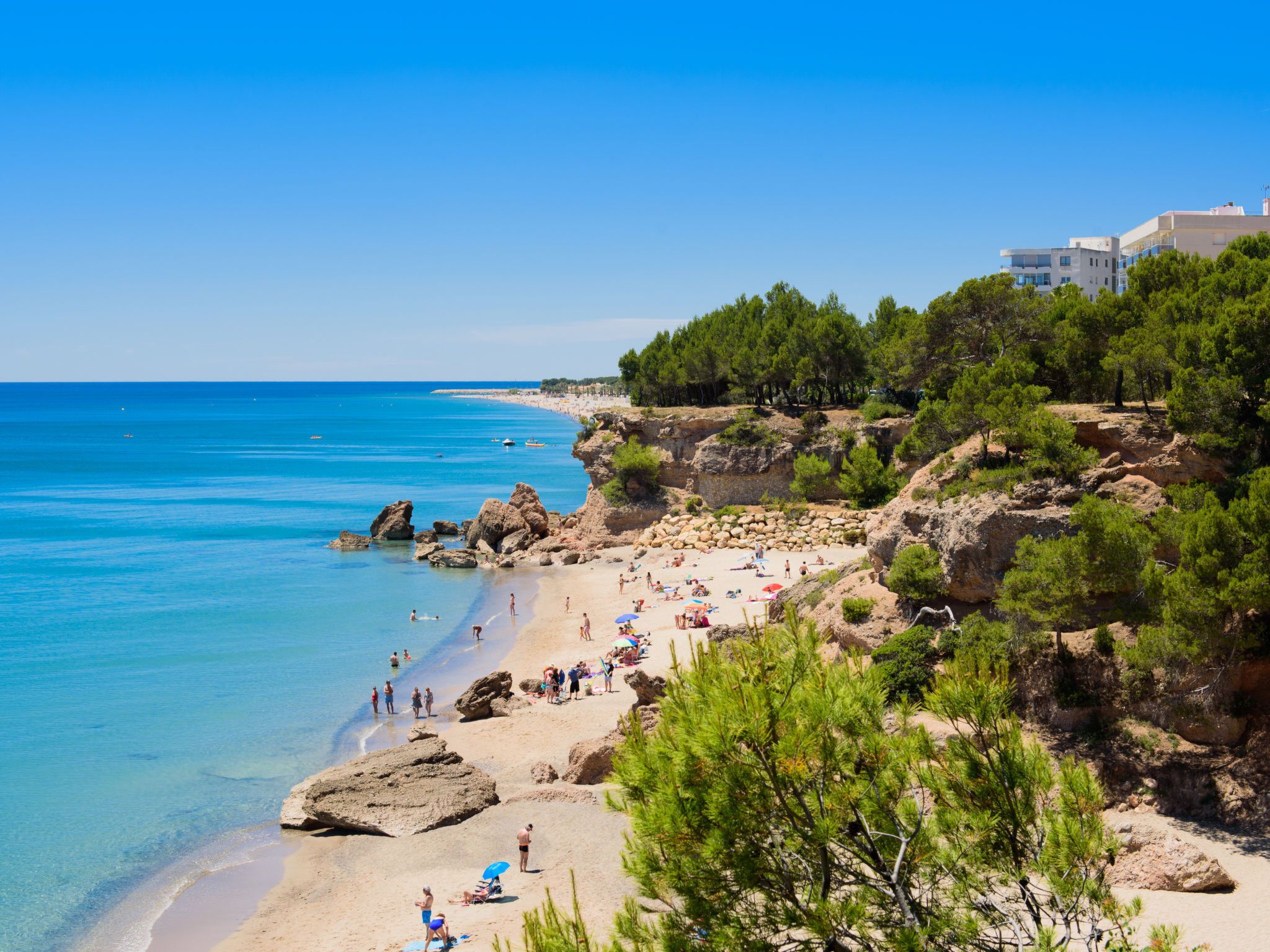 A beach with clear blue waters, rocky terrain, and a few buildings in the background.