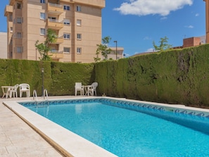 Ein Swimmingpool mit einer gefliesten Kante, umgeben von einer niedrigen Hecke und weißen Stühlen.
