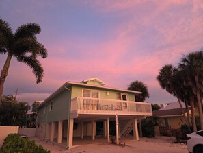 Een huis met twee verdiepingen en een balkon, omgeven door palmbomen, onder een roze lucht.