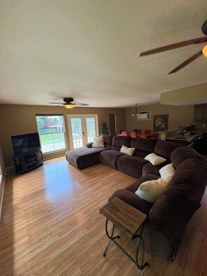 A living room with a sectional sofa, wooden flooring, a ceiling fan, and a TV stand.