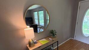 Ein Zimmer mit einem Holzkonsolentisch, einem Spiegel, einer Lampe, einer kleinen Pflanze und einem Fenster.