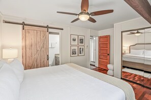 Ein Schlafzimmer mit Bett, Deckenventilator, Holztür und Blick ins Badezimmer.