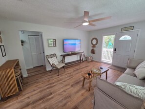 Ein Wohnzimmer mit Deckenventilator, Fernseher, Holzboden, einer Couch und einer zur Außenseite führenden Tür.