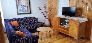 Ein Wohnzimmer mit einer Couch, einem kleinen runden Tisch, einem Schrank mit Fernseher und einer Vase mit Zweigen.