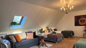 Ein Loft-Zimmer mit schrägverdeckter Decke, integriertem Fenster, einer Chandelierleuchtre, einer Couch, einem Bett und einem kleinen Tisch mit Pflanze.