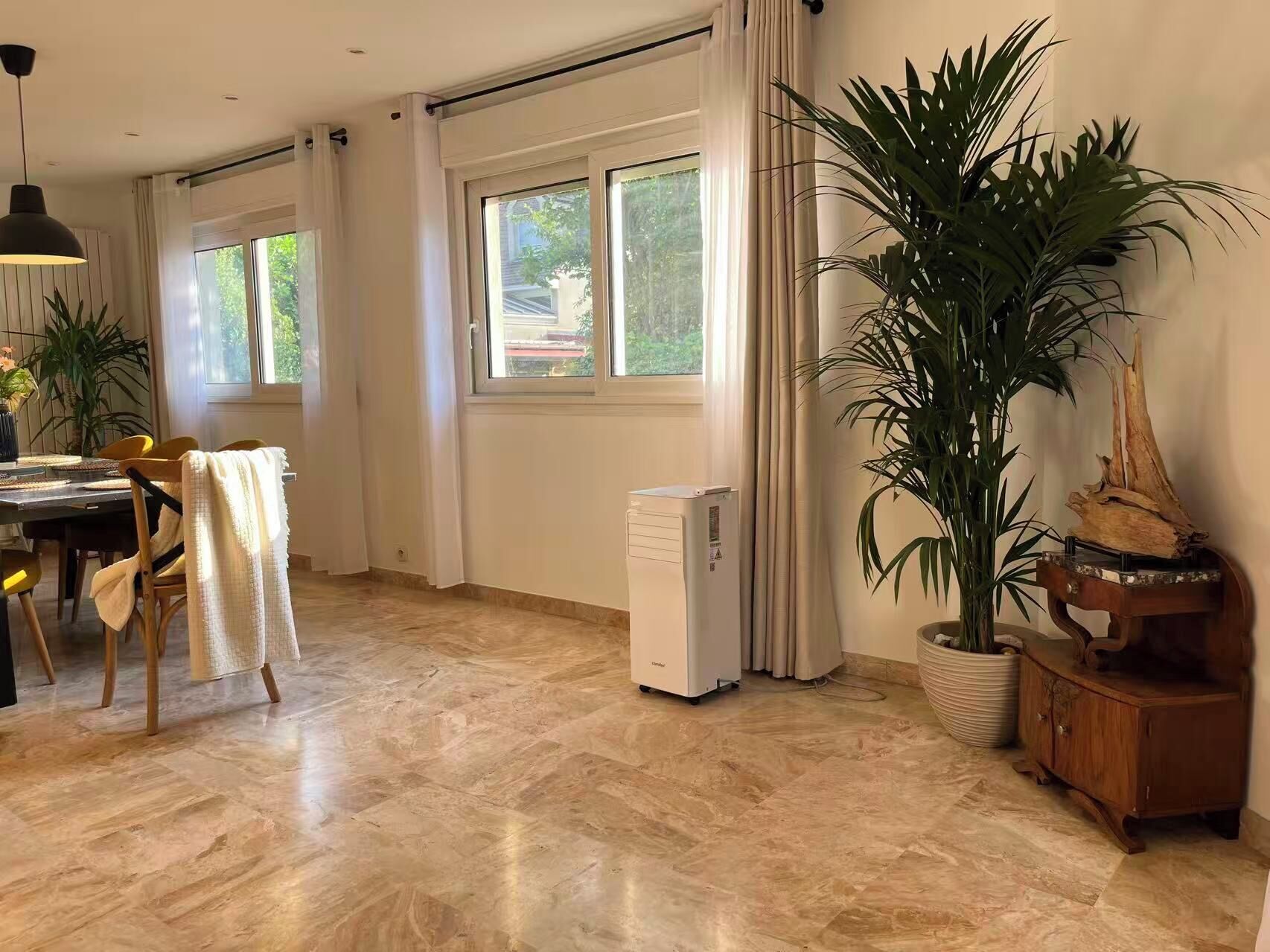 Een ruime kamer met een eethoek, grote ramen, een radiator en een potplant.