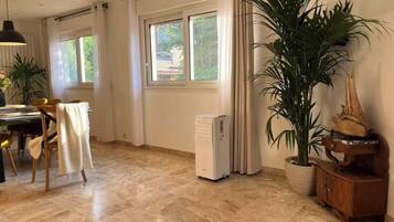 Een ruime kamer met een eethoek, grote ramen, een radiator en een potplant.