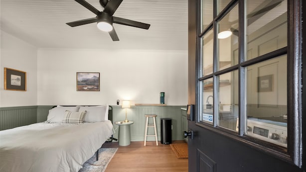 Een slaapkamer met een bed, een plafondventilator, een nachtkastje, een lamp, een kruk en een raam met uitzicht.