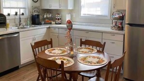 A kitchen with white cabinets, a stainless steel refrigerator, a dining table with floral placemats, and a 'welcome' sign above the windows. - The Mountain Laurel Cottage Near Wildcat ATV Park. (East Bernstadt)