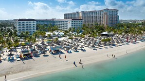 Un hotel frente a la playa con varias construcciones, una playa de arena con sombrillas y palmeras.